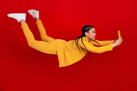 Cheerful woman wearing a yellow suit with cornrow hairstyle taking a selfie against a vibrant red...