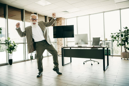 Mature businessman in elegant attire dancing joyfully in a modern office with desk and computerの写真素材