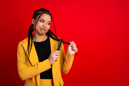 Elegant young woman in a yellow suit with braided hair combing her braids against a vibrant red...