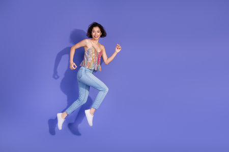 Cheerful woman in fashionable sequined top and casual jeans joyfully posing against a vivid purple...