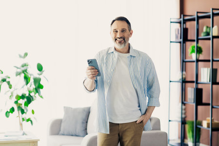 Mature man using smartphone while enjoying a relaxed time in his cozy living space, conveying modern...