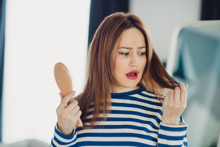 Worried young woman with hairbrush wearing casual striped shirt indoors at home on a bright sunny...