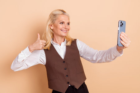 Joyful middle-aged businesswoman giving thumbs up while taking selfie with smartphone against beige backgroundの写真素材
