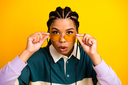Stylish young woman with trendy sunglasses poses against a vibrant yellow background exuding...