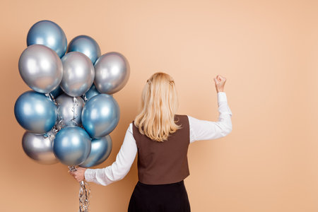 Confident businesswoman with balloons in a celebratory setting on beige backdrop, exuding success...