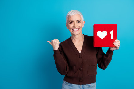 Stylish senior woman holding red like icon block against blue background, showcasing cheerful...