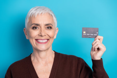 Smiling mature woman holding a credit card in hand against blue background demonstrates finance and...