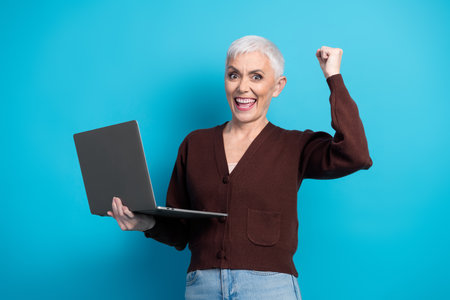 Energetic senior woman with a laptop celebrating success against a bright blue background, showing...