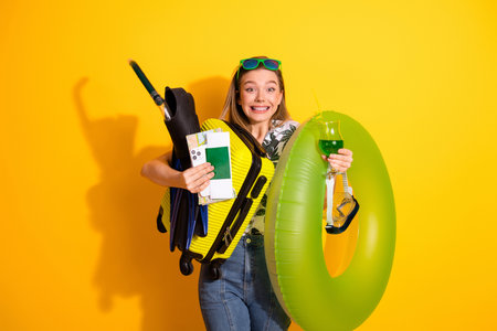 Young cheerful woman holding travel essentials against a vibrant yellow backdrop highlighting her...