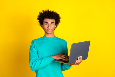Young man holding a laptop against a vibrant yellow background showcasing casual style and a teal...