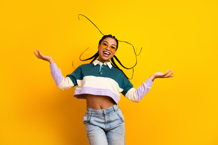 Cheerful young woman with braided hair poses playfully against a vibrant yellow backdrop, radiating...