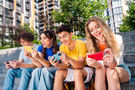 Group of cheerful young friends enjoying a sunny day, engaging with smartphones on urban outdoor...