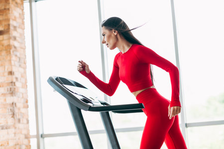 Fitness trainer sporty woman in red activewear practicing cardio on treadmill in a stylish gym interiorの写真素材