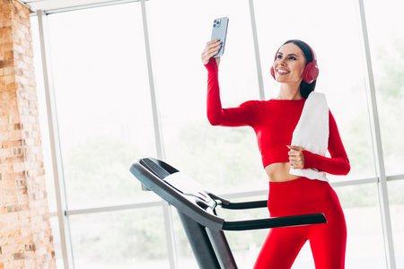 Young woman in red activewear smiling while exercising on a treadmill and taking a selfie at a...
