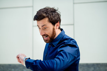 Young businessman checking time on wristwatch in urban outdoor setting while expressing concern and...
