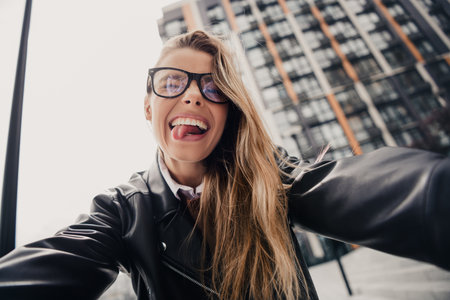 Young stylish woman taking a cheerful selfie outdoors on a city street during daylight with urban...