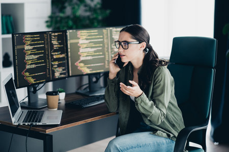 Young female programmer working on coding tasks at her desk in a modern workspace with multiple...