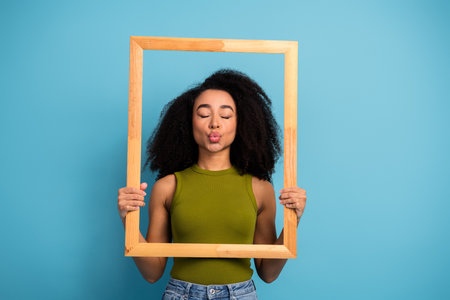 Woman in casual green top holding wooden frame and playfully blowing a kiss against a blue...