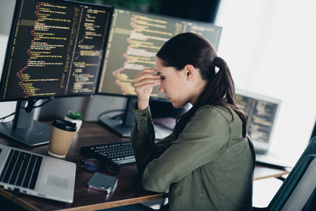 Young female programmer stressed over coding issue at home workspace with multiple monitors showing...