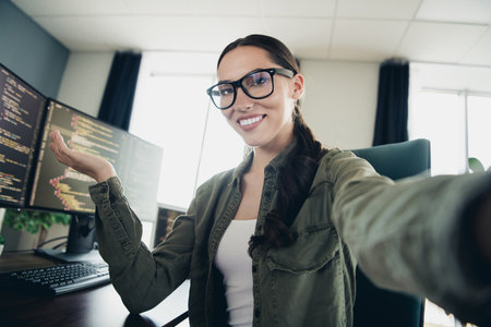 Young female programmer at her workspace holding a welcoming gesture, coding on multiple monitors,...