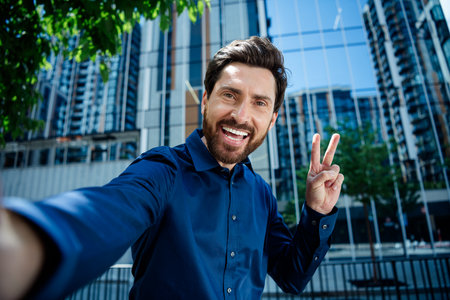 Confident young businessman taking a selfie outdoors in a vibrant urban setting with modern office...
