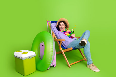 Young man lying on a deck chair with holiday props enjoying a tropical beverage against a vibrant green backdropの写真素材