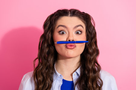 Playful young woman balancing a pen on her upper lip in front of a vibrant pink background, showcasing charmの写真素材