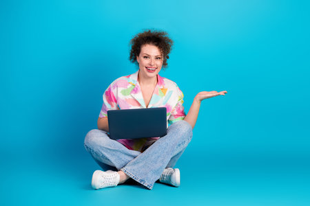 Young smiling woman seated cross-legged holding a laptop, gesturing with hand, against a vibrant blue backgroundの写真素材