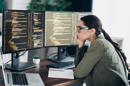 Focused female software developer analyzing code on multiple monitors in modern home office environmentの写真素材