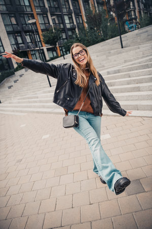 Young woman joyfully posing outdoors in casual chic outfit including leather jacket and denim, exuding happinessの写真素材