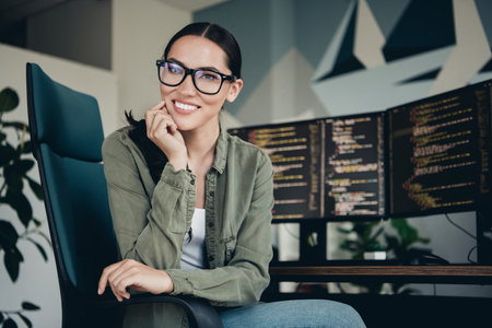 Confident young female programmer at modern workspace with computer setup coding and smiling confidently indoorsの写真素材
