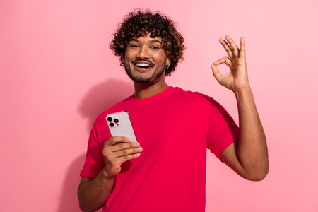Cheerful young man holding smartphone and making ok sign against pink background, expressing positivity and easeの写真素材