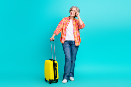 Senior woman with yellow suitcase and glasses smiling on teal background, demonstrating leisure and travel lifestyleの写真素材