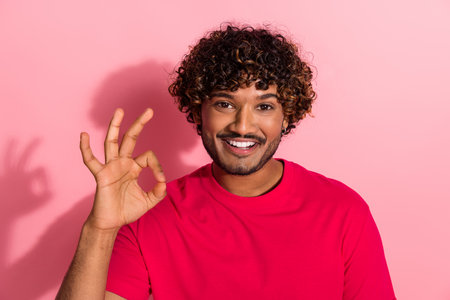 Happy young man in a red t-shirt gestures okay and smiles against a pink background with a cheerful expressionの写真素材