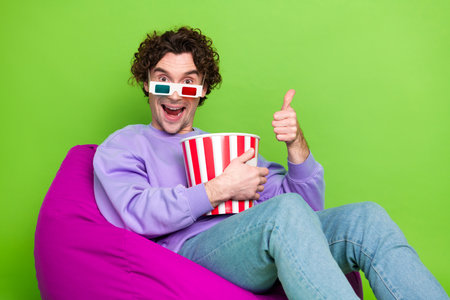 Young man in 3D glasses enjoying popcorn while seated on a bean bag showing thumbs up against green backgroundの写真素材