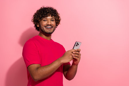 Young man in a red t-shirt holding a smartphone against a pink background, expressing positivity and modern lifestyle.の写真素材