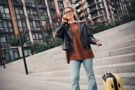 Lovely Attractive Young Woman with Luggage Listening to Music Outdoors in Urban Settingの写真素材