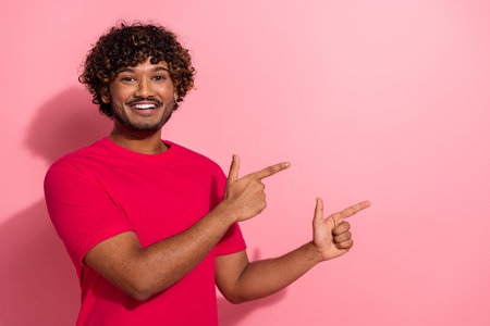 Cheerful young man in red t-shirt making pointing hand gestures against a vibrant pink backdropの写真素材