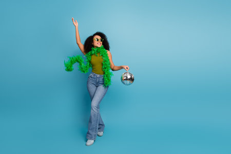 Fashionable young woman in trendy outfit holding a disco ball and gesturing joyfully against a...