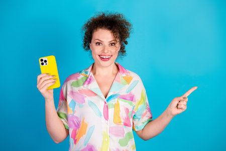 Cheerful woman in colorful shirt holding yellow phone and pointing, happy with vibrant blue...