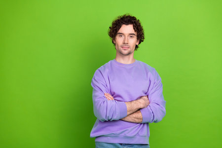 Young curly-haired man wearing casual purple sweatshirt posing confidently with arms crossed against...