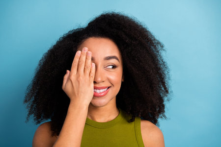 Smiling young woman with curly hair covering one eye playfully against blue background in casual...