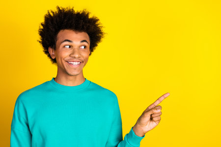 Cheerful young man in a teal sweatshirt pointing to the side against a bright yellow background with...