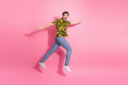 Excited young man in a vibrant yellow tropical shirt and jeans happily leaping against a bright pink...