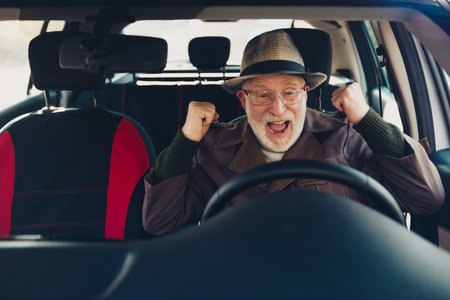 Happy elderly man cheering with excitement while sitting in a car on a sunny day, enjoying the new...