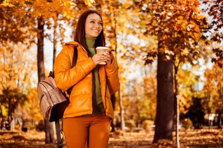Smiling woman in autumn attire enjoys a coffee while walking in a colorful fall forest with vibrant...