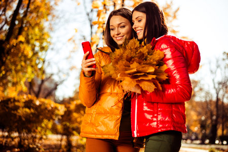 Two young women enjoying a sunny autumn day in the park, smiling and holding a bouquet of vibrant...