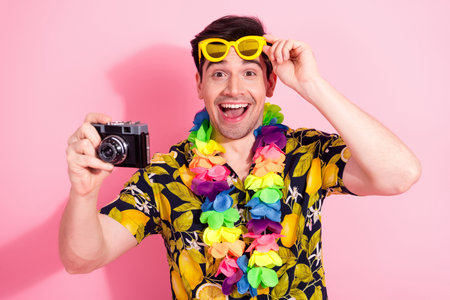 Excited young man with yellow sunglass and tropical attire taking a photo with a vintage camera on...