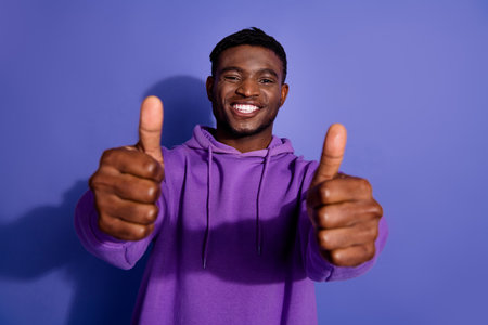Smiling young man in a purple hoodie giving thumbs up on a vibrant violet background, showing...