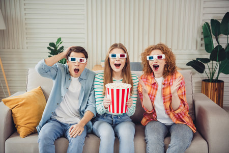 Group of three young friends watching a 3D movie at home with popcorn, expressing surprise and joy...